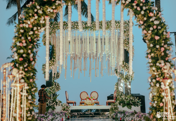 Wedding stage on the beach with a floral arch, macrame canopy, and mirrored base at twilight.