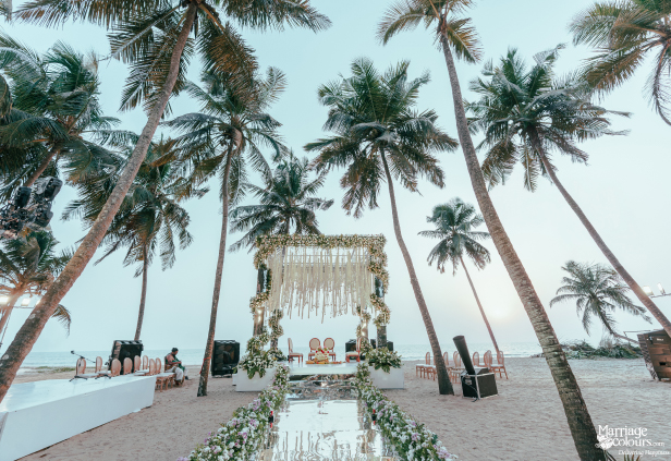 Scenic beach wedding mandap with mirrored aisle, macrame canopy, and floral decor under palm trees.