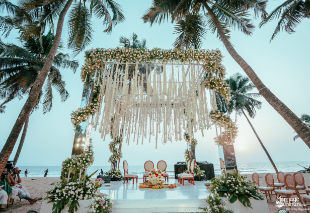 Wedding mandap on the beach with mirrored pillars, white floral garlands, and macrame canopy at sunset.