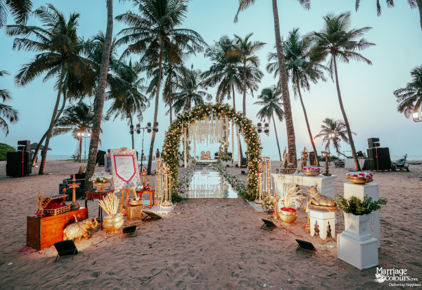 Elaborate beach wedding entrance with floral arch, mirrored aisle, and gold traditional props under palm trees.