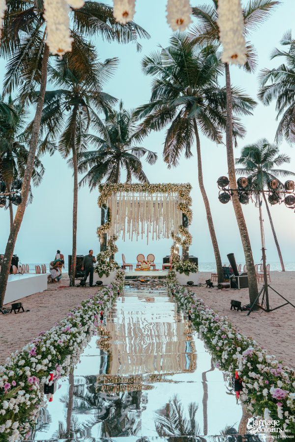 Mirrored aisle leading to a white macrame and floral wedding mandap under tall palm trees.
