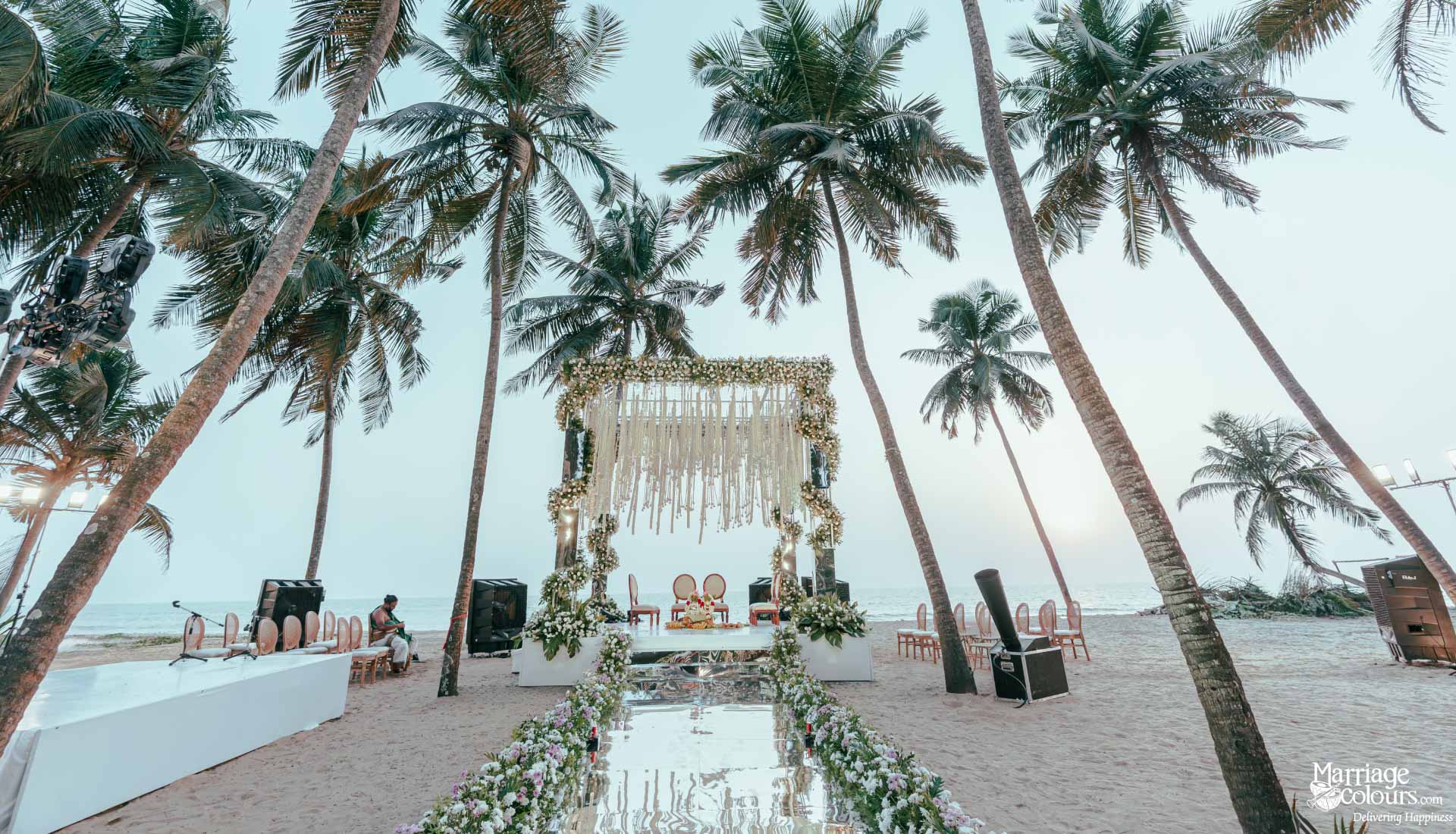 Scenic beach wedding mandap with white macrame, floral decor, and mirrored aisle under palm trees.