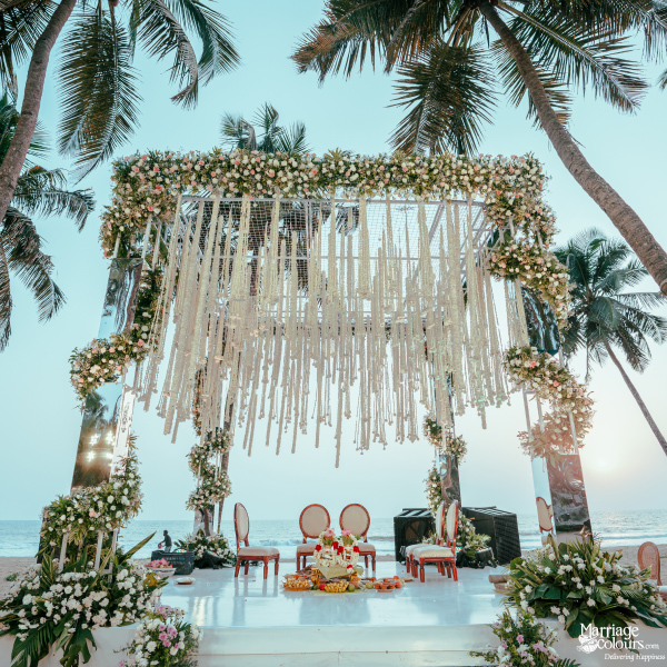 Beach wedding mandap with mirrored pillars, white macrame, and lush florals under tall palm trees.