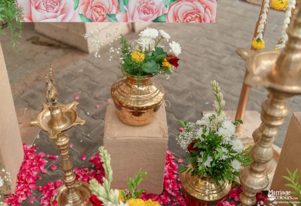 Traditional wedding ritual items: brass kalash pots with flowers, lamps, and pink rose petals on the ground.