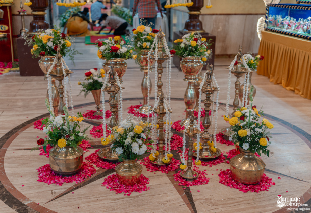 Traditional Hindu ritual setup with brass oil lamps, kalash pots, and pink rose petals on the floor.