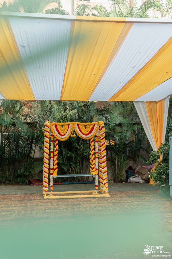 Outdoor wedding swing decorated with red, yellow, and white marigold garlands under a striped awning.