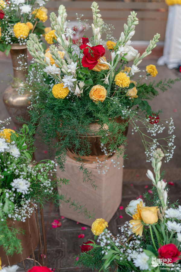 A bronze vase with vibrant wedding flowers, including red roses, yellow marigolds, and tuberoses.
