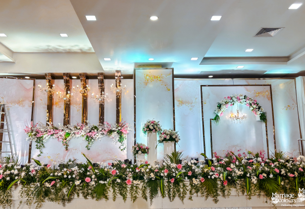 An elegant wedding reception stage is decorated with floral arrangements, a large archway, and white and gold backdrops.