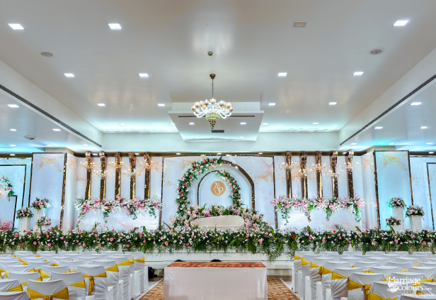 An elegant wedding reception hall with a grand stage decorated in white and gold, featuring a central floral arch and rows of guest chairs.