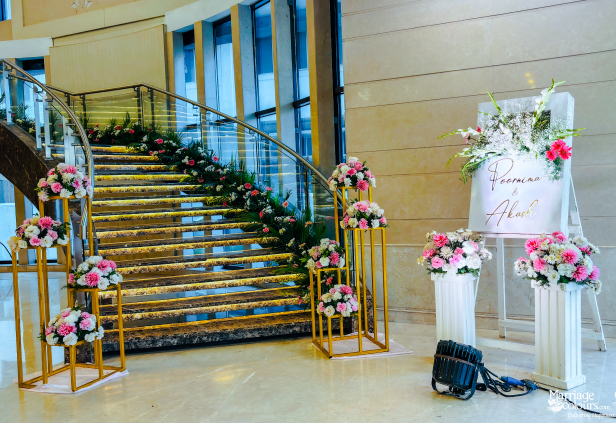An elegant staircase decorated with floral arrangements leads to a wedding reception welcome sign with a sign and more flowers on stands.