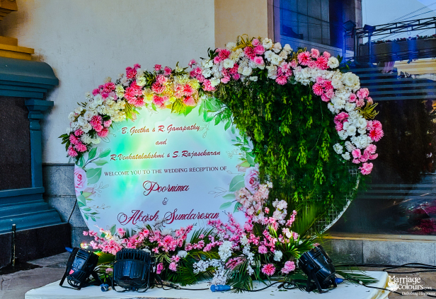 A circular welcome sign for a wedding reception is decorated with a vibrant arch of pink and white flowers and green foliage.