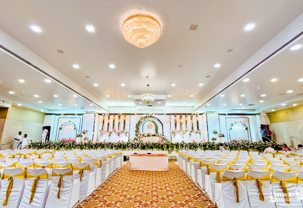 A grand wedding reception hall with a decorated stage, a central aisle with an ornate runner, and rows of chairs with gold sashes.