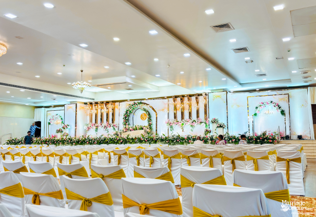 A wedding hall with rows of white chairs facing a beautifully decorated stage with a floral arch and elegant backdrop.