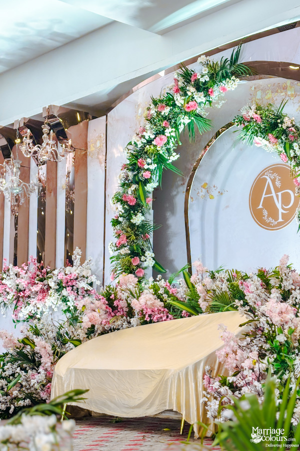 A close-up of a wedding stage with a white sofa, a large floral arch, and a backdrop featuring a gold frame with the initials 'AP'.