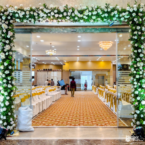 An ornate floral arch frames the glass entrance to a wedding reception hall, showing a view of the aisle and guest seating inside.