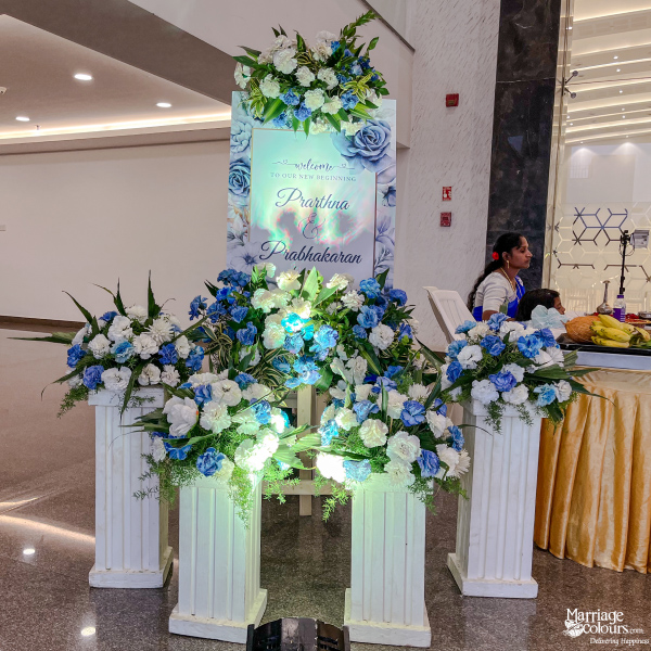 Welcome sign for Prarthna & Prabhakaran featuring blue flowers on white Roman-style pedestals.