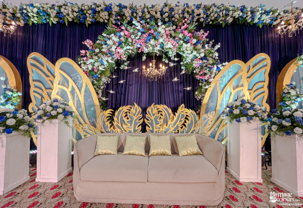 Wedding stage with blue drapes, golden butterfly wings, a central sofa, and lush floral arch.