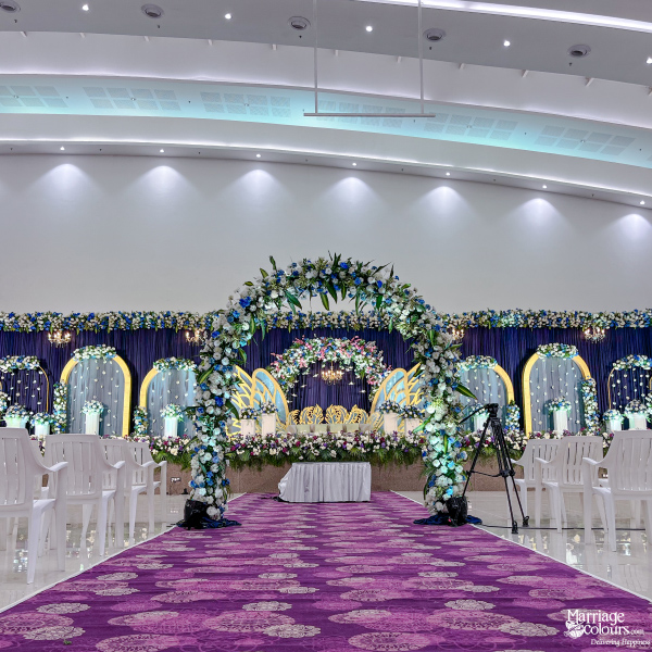 Grand wedding hall stage with a floral arch, purple aisle, and rows of white plastic chairs.