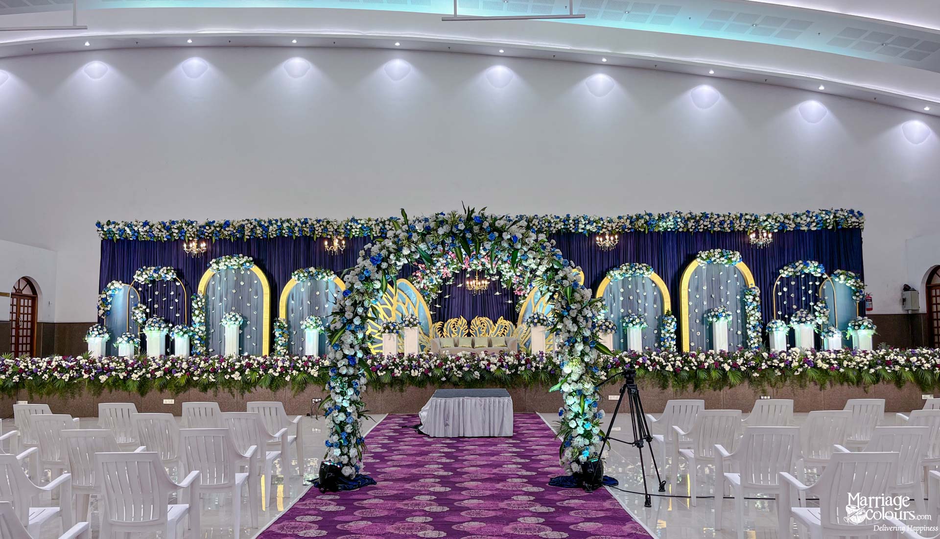 Grand indoor wedding stage featuring a floral arch, royal blue backdrop, and white guest seating