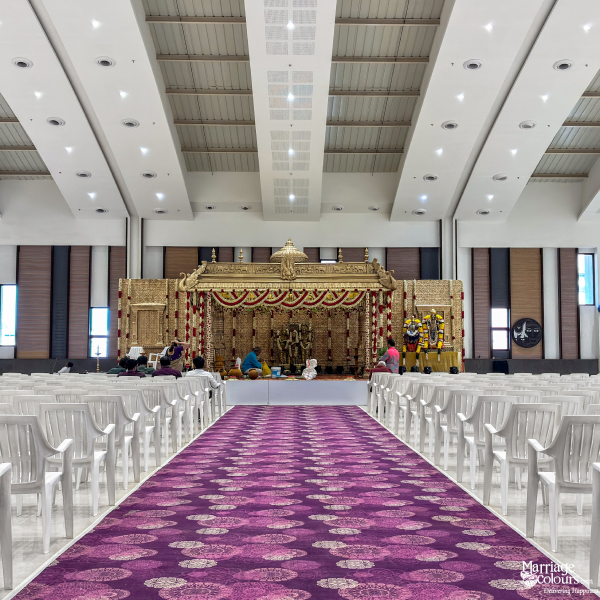Wide view of a golden South Indian stage, purple aisle, and rows of white plastic chairs in a hall.