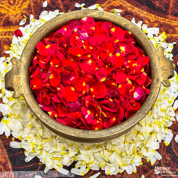 Close-up of a brass pot filled with red rose petals, surrounded by white and yellow petals.