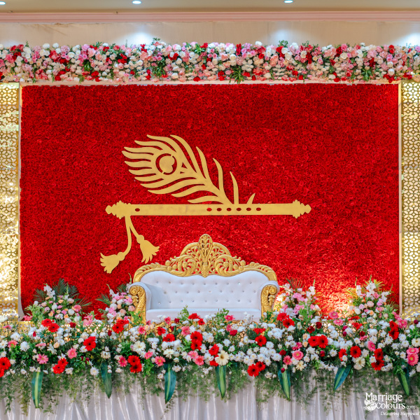 Vibrant wedding stage with a bold red floral wall, white and gold sofa, and golden flute and feather motif.