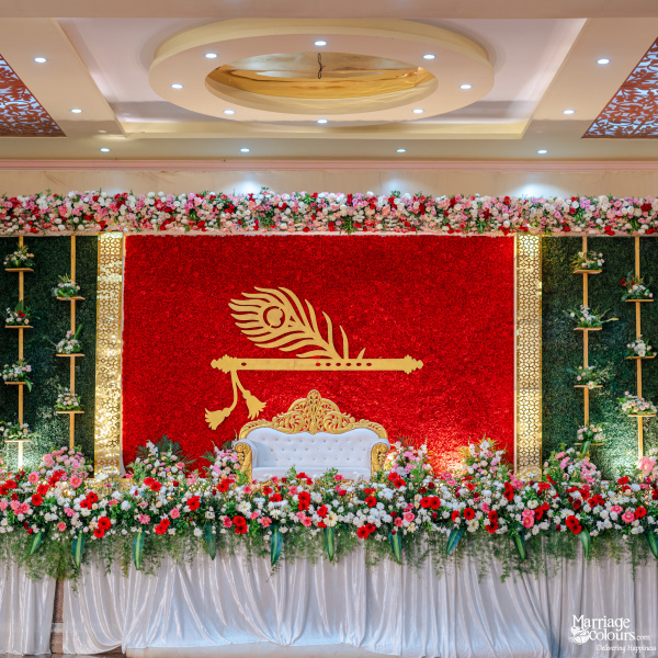 Vibrant wedding stage with a red floral wall, white and gold sofa, and Lord Krishna's flute motif.