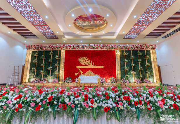 Grand wedding stage with a striking red floral wall, white and gold sofa, and green foliage panels.
