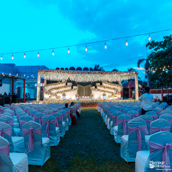 Outdoor evening wedding with a flower-covered stage, rows of white chairs, and pink sashes.