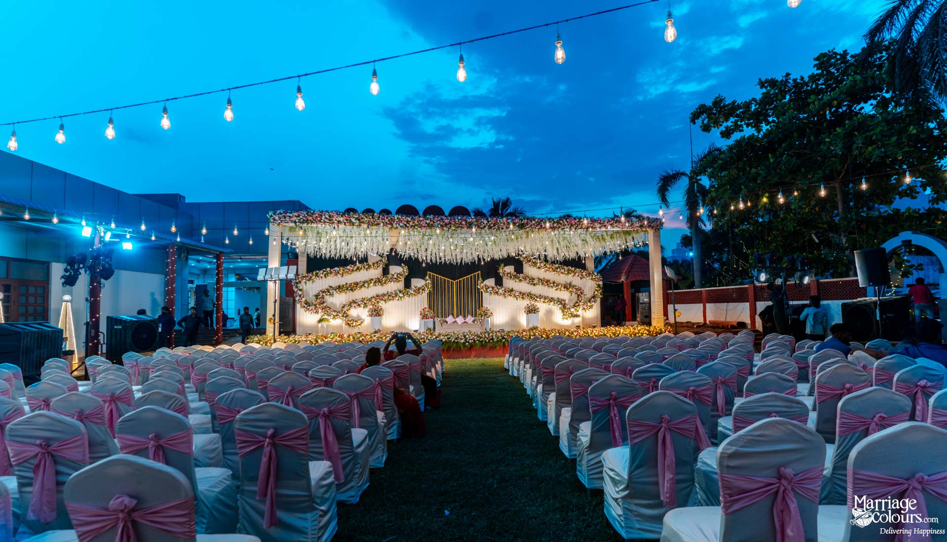 Outdoor evening wedding setting with a floral stage, rows of chairs, and hanging string lights.