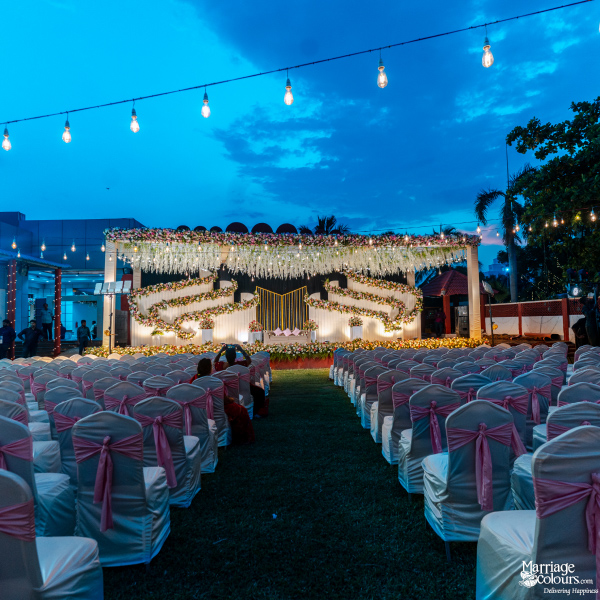Outdoor evening wedding ceremony setup with white chairs, pink sashes, and a tiered floral stage.