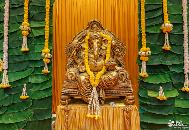 Close-up of a golden Ganesha idol on a throne, garlanded with marigolds, against a banana leaf backdrop.