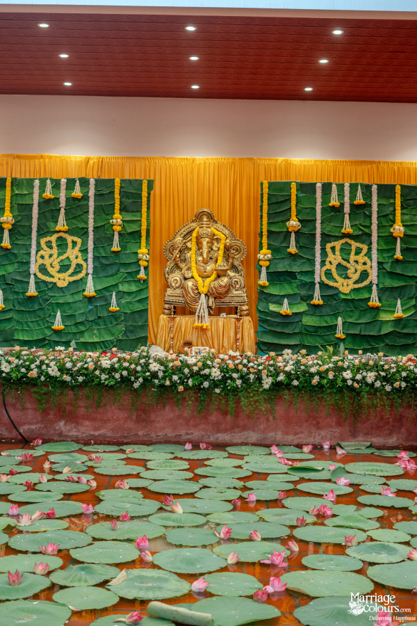 Traditional wedding stage with a golden Ganesha idol, banana leaf backdrop, and a lotus pond foreground.