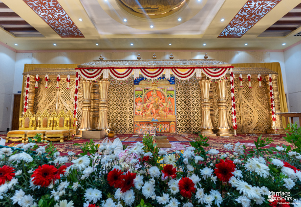 Grand wedding stage with a central Hindu deity painting, ornate gold carvings, and red and white flowers.