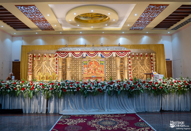 Grand wedding stage with a central Hindu deity painting, ornate gold carvings, and red and white flowers.