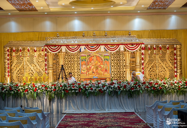Grand wedding stage with a central Hindu deity painting, ornate gold carvings, and a lush floral border.