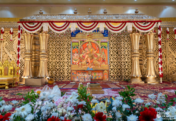 Grand traditional wedding stage with a central Hindu deity painting, ornate gold carvings, and a lush floral border.