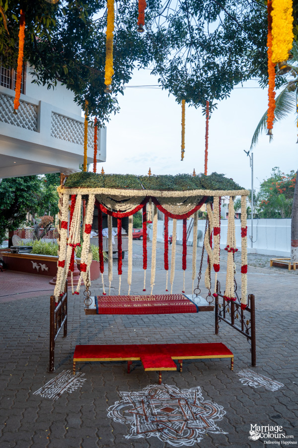Traditional wedding swing decorated with white, red, and green garlands, set up outside a house with a kolam.