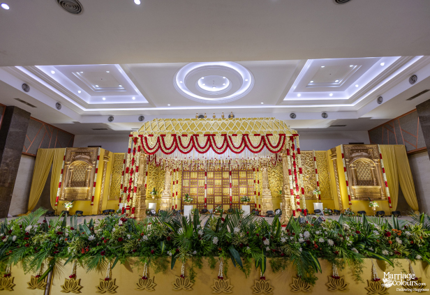 Grand traditional wedding stage with a gold mandap, intricate gold carvings, and a lush green and white floral foreground.