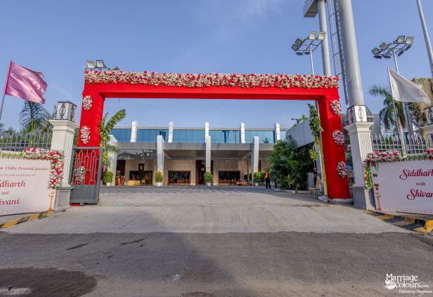 Wedding entrance arch in bright red, decorated with white and red flowers, leading to a modern building.