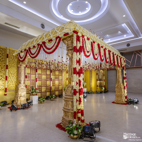 Grand golden mandap with ornate pillars, covered in red and white flower garlands, against a gold lattice backdrop.