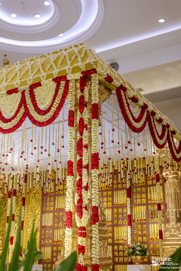 Close-up of a wedding mandap with white and red garlands, hanging white tassels, and a gold lattice background.