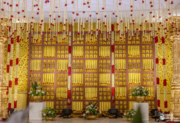Wedding backdrop with a golden lattice pattern, white and red hanging garlands, and floral arrangements.