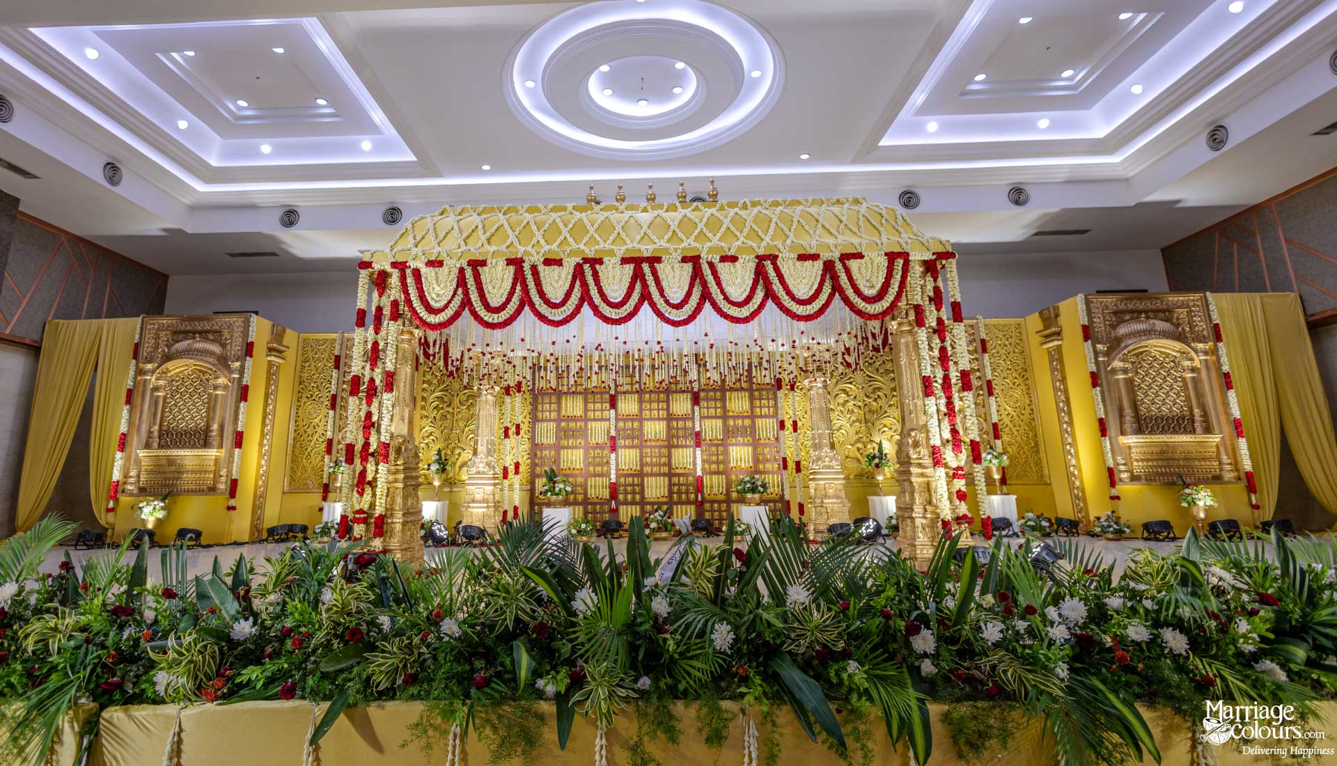 Grand, ornate wedding stage with a gold arch, red and white floral garlands, and lush green foreground plants.