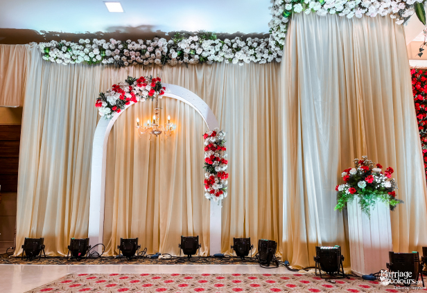 Wedding stage with cream drapery backdrop, a white arch, chandelier, and red and white flowers.