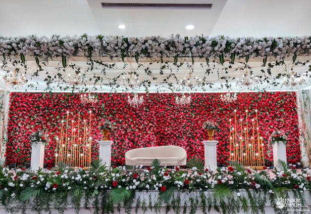 Wedding stage with red flower wall, white sofa, chandeliers, and lush white and green floral border.