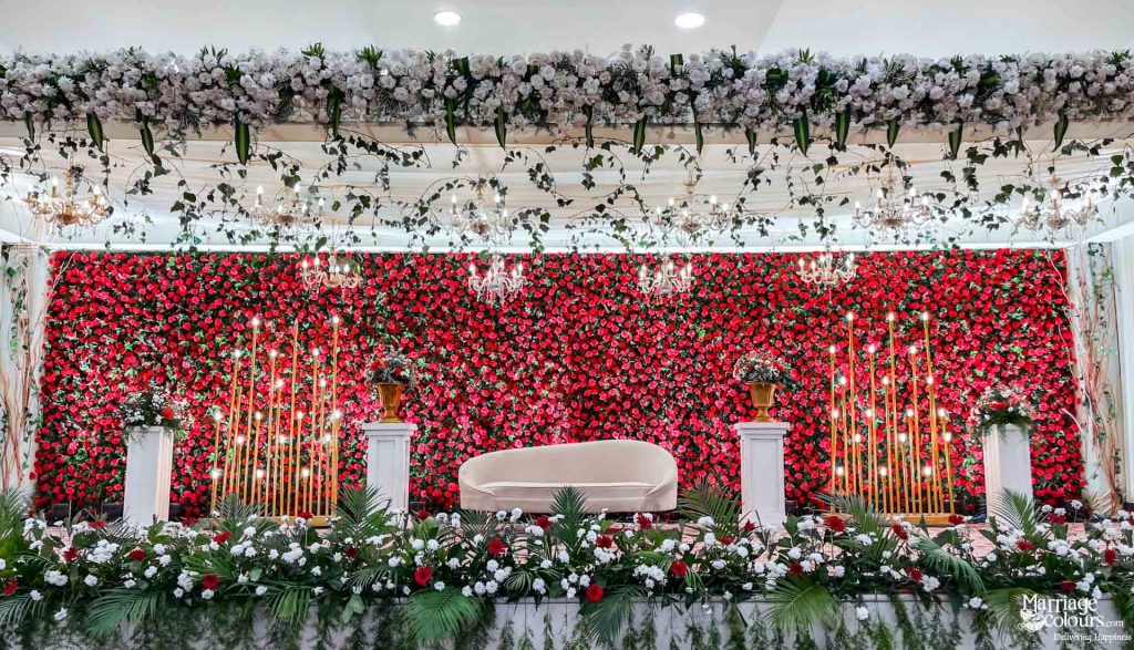 Wedding stage with red flower wall, white curved sofa, and lush white and green floral border.