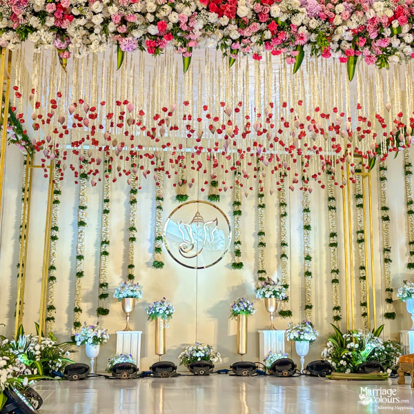 Ornate wedding stage with Ganesha centerpiece, golden pillars, floral strings, and a large pink and white flower arch.