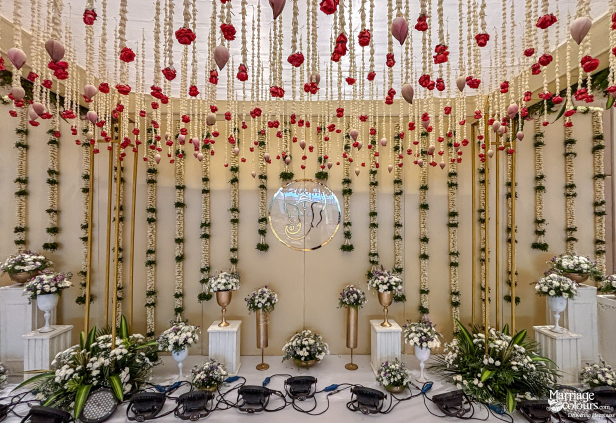 Detailed wedding stage with hanging red roses, white strings, Ganesha centerpiece, and lush floral arrangements.