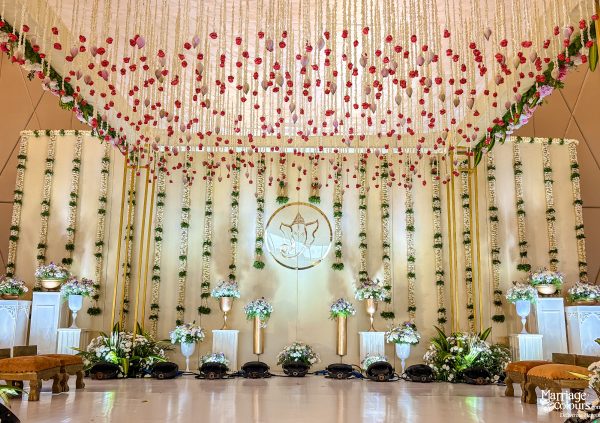Lavish wedding mandap with a hanging floral canopy, Ganesha emblem, white cloth backdrop, and golden accents.